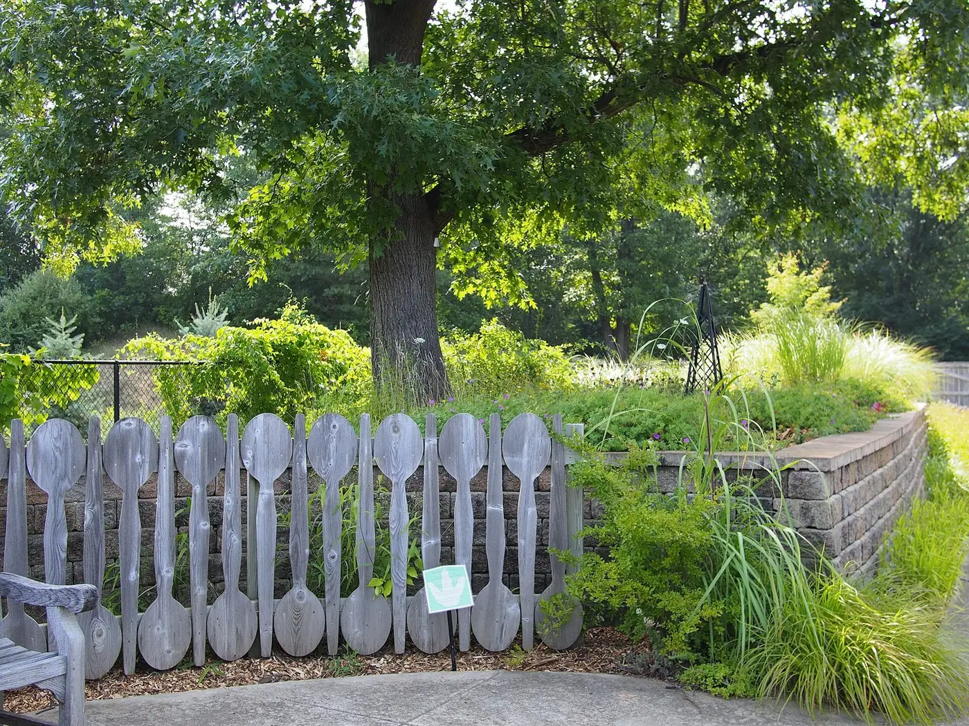 Garden at Leila Arboretum with a large shade tree behind a fence of oversized wooden spoons
